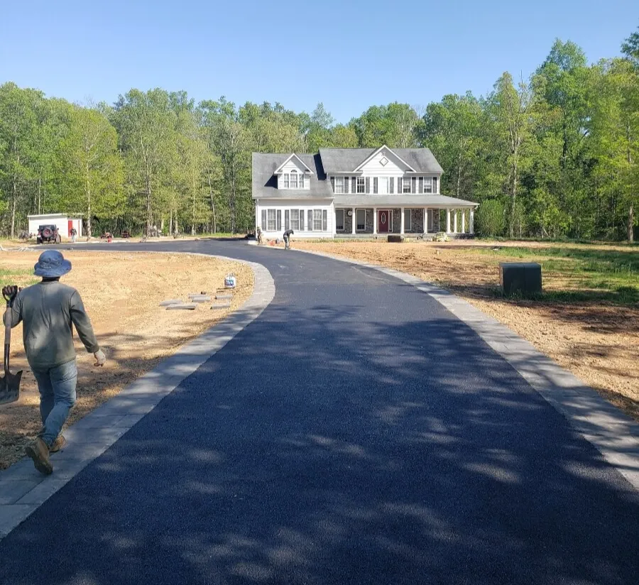 A worker walks along a newly paved driveway leading to a large house surrounded by trees.