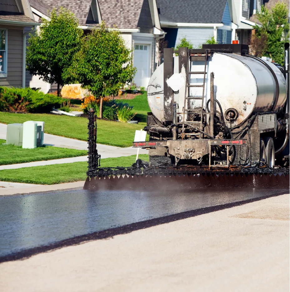 sealcoating-2 A truck sprays fresh asphalt onto a suburban street in front of houses and green lawns.