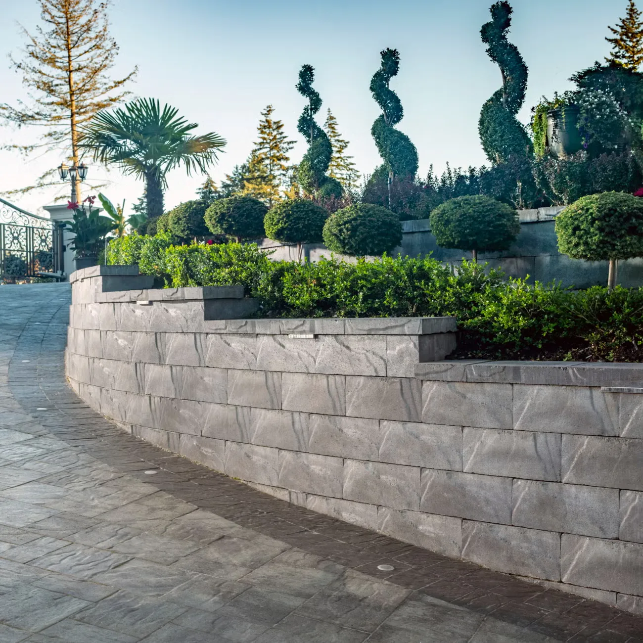 Tiered stone retaining wall with manicured shrubs and spiral-shaped topiary plants in a landscaped garden.