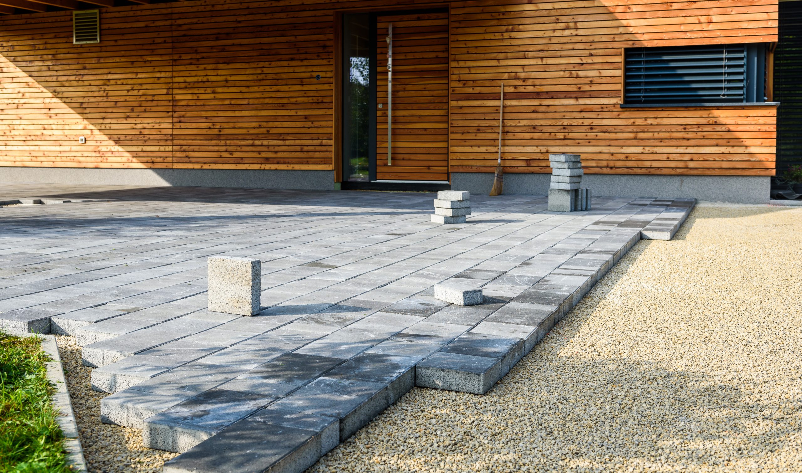 Gray paving stones being laid in front of a modern wooden house, with some stones stacked nearby.