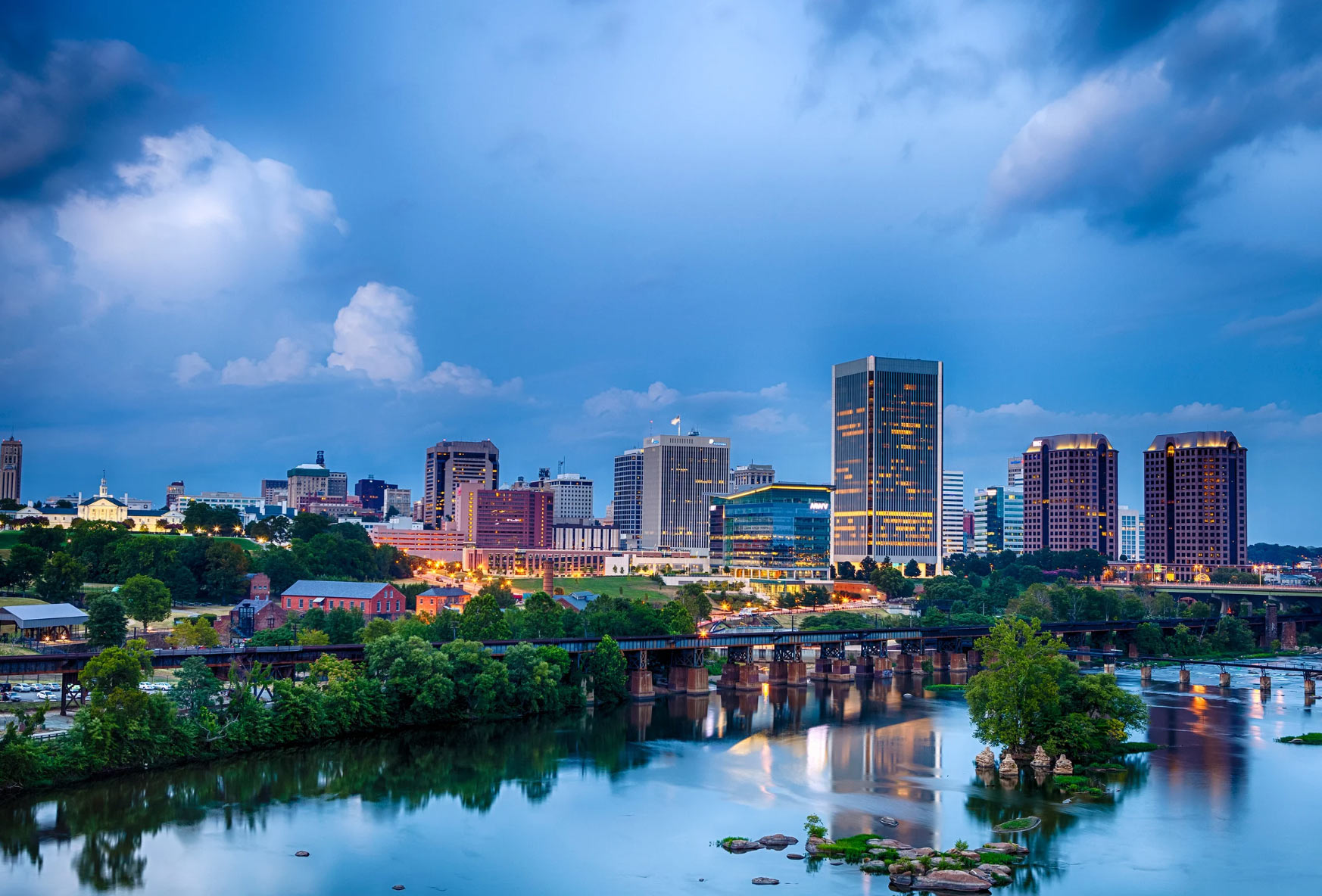 virginia-img City skyline at dusk with illuminated buildings reflected in a river, trees in the foreground, and a cloudy sky.