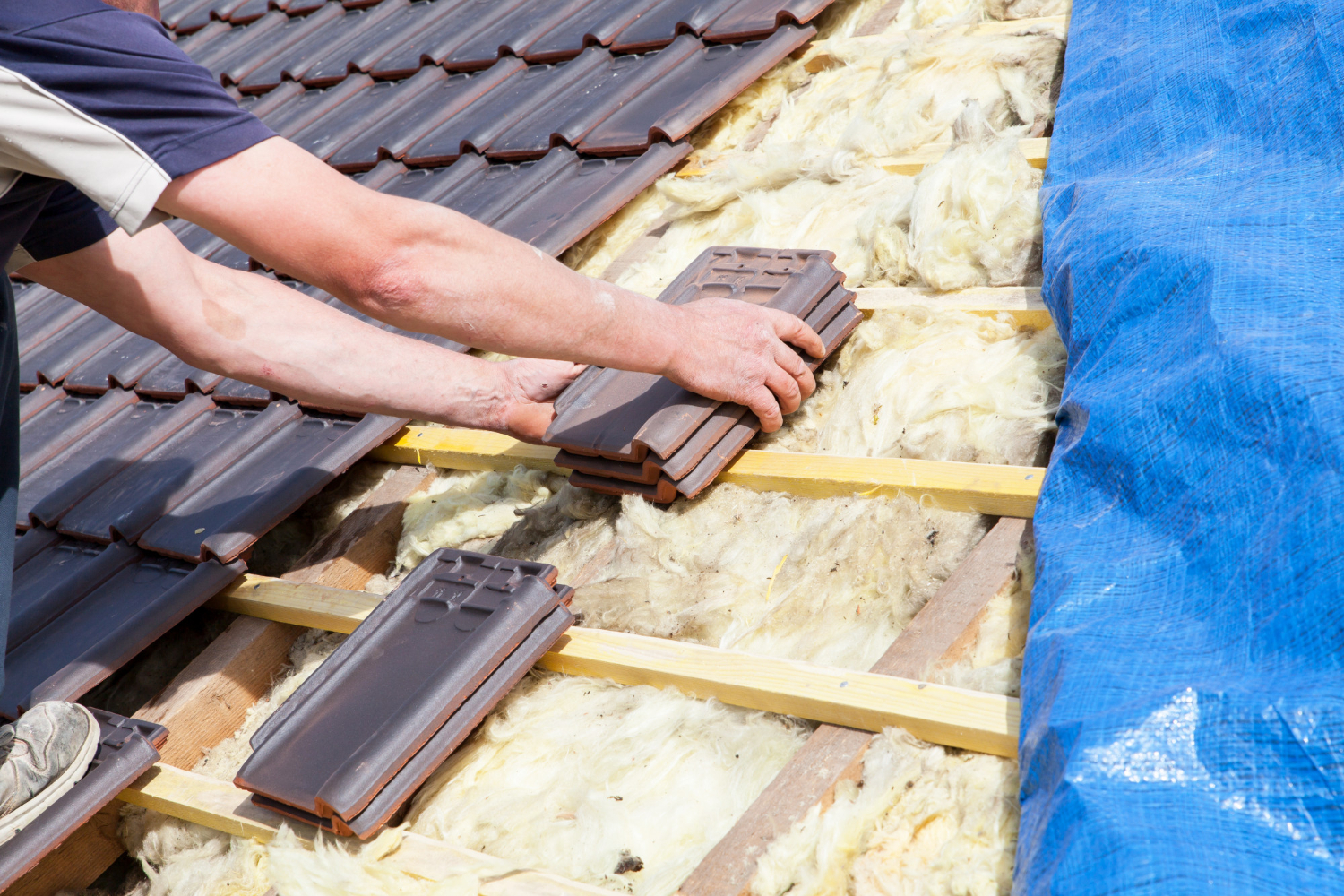 A person installing brown roof tiles over yellow insulation and wooden battens on a roof.