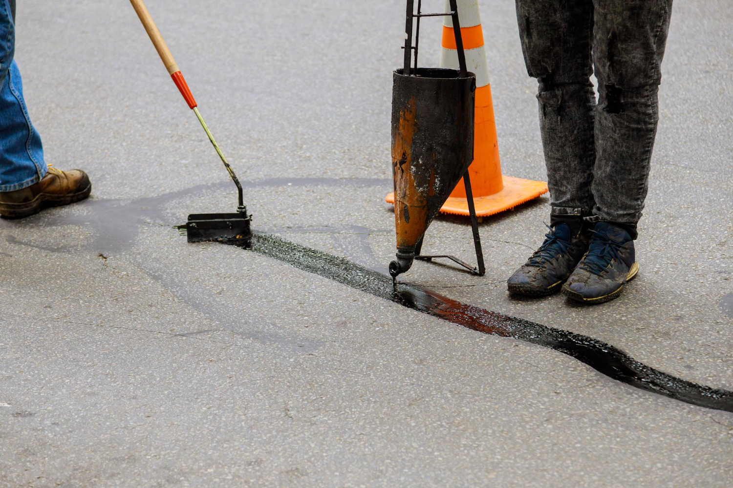 21460467 Workers sealing a crack in asphalt with hot tar, using tools next to an orange traffic cone.